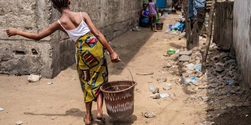 A young girl balances a water bucket through a sandy alleyway. With no indoor plumbing, daily water collection is part of life.